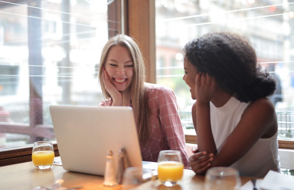 Two young women are smiling and sharing a laugh as they watch a video together on a laptop in a brightly lit cafe, with glasses of orange juice on the table.